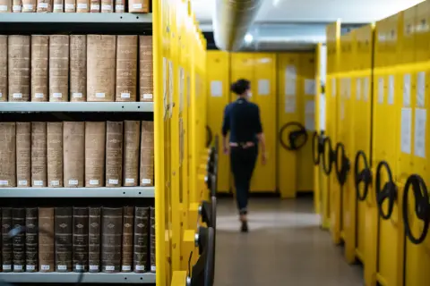 Rayons de livres anciens dans les archives départementales d'Indre-et-Loire en ligne, une personne se déplace dans l'allée.