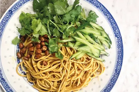 En läcker asiatisk nudelsallad med krispig gurka, rostade jordnötter och färsk koriander på en vackert mönstrad tallrik.