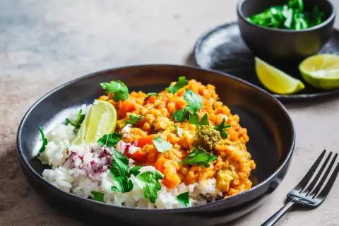 Un plat de riz blanc garni d'un curry de lentilles corail et légumes, parsemé de coriandre fraîche et de quartiers de citron vert. Une recette d'été parfaite.