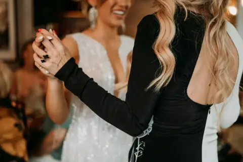 Deux femmes dansent, l'une en robe blanche scintillante, l'autre en robe noire. Elles se tiennent la main, un geste pour faire honneur au mariage.