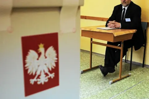 Mąż zaufania w garniturze siedzi przy biurku w lokalu wyborczym, obok urny z godłem Polski.