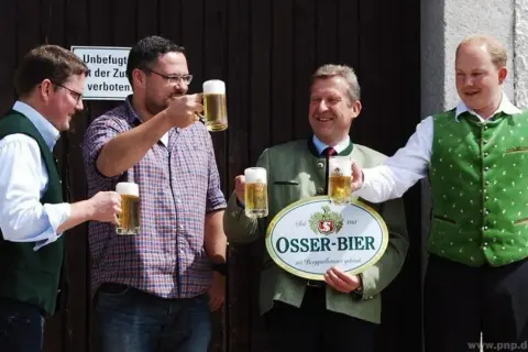 Vier Männer stoßen mit Biergläsern an. Einer hält ein Schild der Osser Brauerei.
