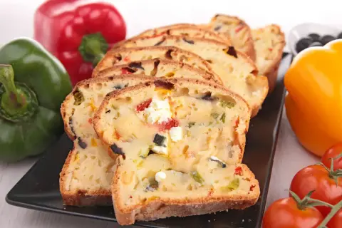 Tranches d'un cake savoureux aux olives et feta, entouré de poivrons colorés et de tomates.