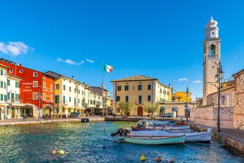 Colorful buildings line a harbor with boats, an Italian flag, and a bell tower under a clear blue sky.