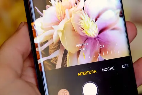 Mano sujetando móvil, mostrando interfaz de cámara con flores y ajustes de apertura. Aprende como hacer un selfie con el móvil.