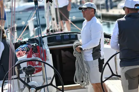 Equipaggio barca a vela pronto per la regata. Un uomo tiene una cima, una donna è seduta, mentre un altro uomo è al timone.