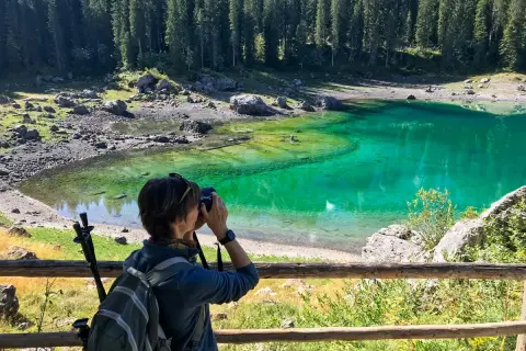 Donna con zaino e macchina fotografica cattura la bellezza del Lago di Carezza, un luogo imperdibile tra le meraviglie del Trentino Alto Adige cosa vedere.