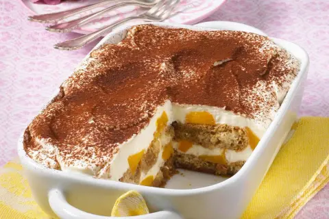 Ein cremiges Pfirsich Tiramisu, bestäubt mit Kakao, in einer weißen Auflaufform. Saftige Pfirsichstücke und Löffelbiskuits sind geschichtet.