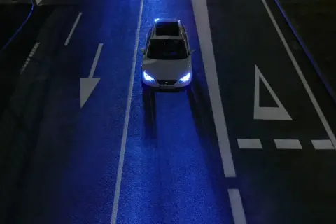 Un coche blanco con las luces encendidas, conduciendo de noche por una carretera oscura con marcas viales blancas y un reflejo azulado.