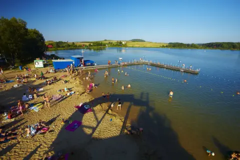 Słoneczny dzień nad zalewem w Siamoszycach. Ludzie odpoczywają na plaży i kąpią się w wodzie. Sprawdź cennik atrakcji!