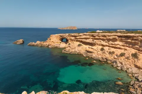 Costa rocosa con una cueva natural en Cala Comte, aguas turquesas y el mar azul profundo.