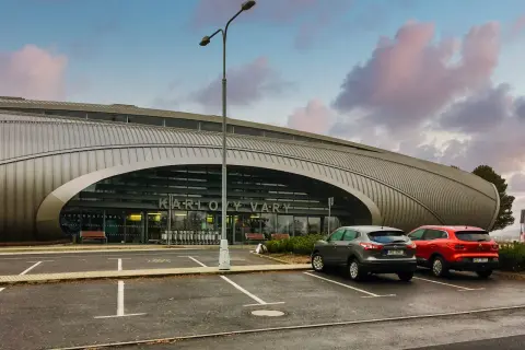 Nowoczesne lotnisko w Czechach, Karlowe Wary, z charakterystyczną, falistą fasadą. Dwa samochody stoją na parkingu przed wejściem.