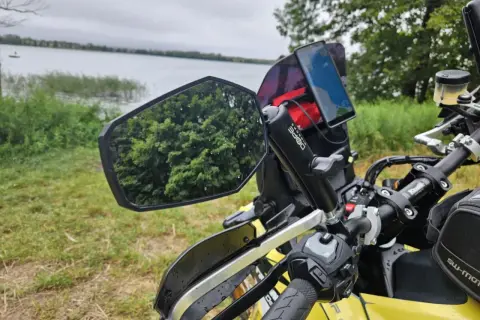Lusterko motocyklowe z odbiciem drzew i telefonem nawigacyjnym na kierownicy, nad jeziorem.
