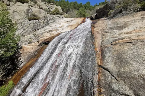 El agua desciende por la roca en la cascada del Hornillo, rodeada de vegetación y rocas graníticas bajo un cielo azul.