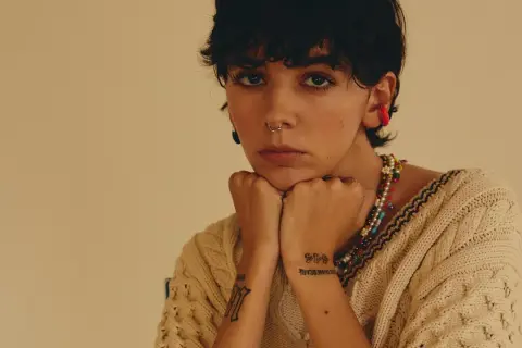 Un'ariete cantante con piercing e tatuaggi, indossa un maglione beige e una collana colorata.