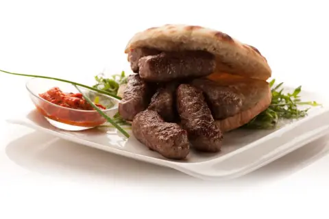En tallrik med balkan mat: saftiga köttfärsrullar i pitabröd, serverade med en röd röra och grönsaker.
