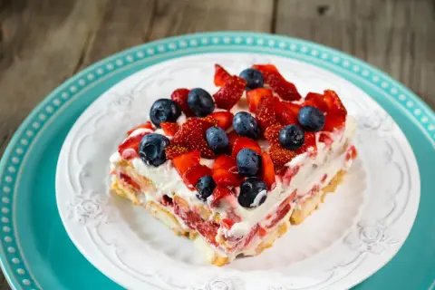 Tranche de tiramisu aux fruits rouges, fraises et myrtilles, sur une assiette blanche ornée, posée sur une assiette turquoise.