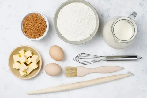 Ingredientes para hornear: harina, azúcar moreno, mantequilla en cubos, huevos, leche en jarra, batidor, brocha y rodillo de madera.