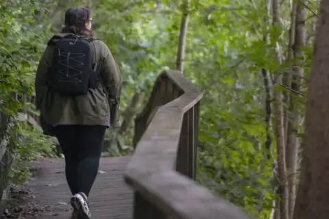 En person med ryggsäck vandrar på en träbro genom en lummig skog. En härlig vandring i Dalarna.
