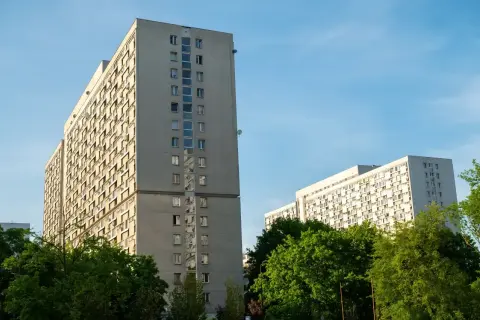 Dwa wysokie, szare bloki mieszkalne z balkonami, otoczone zielenią. W tle błękitne niebo. Taki widok to codzienność wielu lokatorów, którzy co miesiąc płacą **czynsz**.