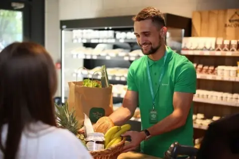 Sprzedawca w zielonej koszulce podaje klientce koszyk z zakupami w sklepie spożywczym.