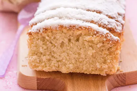 Bizcocho de coco esponjoso, cubierto con coco rallado, servido en tabla de madera.