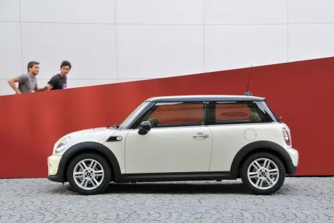 Biały Mini Cooper r56 z czarnym dachem stoi na bruku. Dwóch mężczyzn biegnie za nim na tle czerwono-białej ściany.