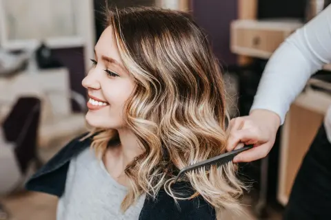 Mujer sonríe mientras estilista peina su cabello con tinte sobre mechas rubias, creando un look radiante.