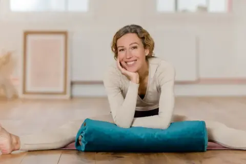 Frau lächelt in einem hellen Raum, Beine gespreizt, mit einem blauen Bolster für Nadi Yoga.