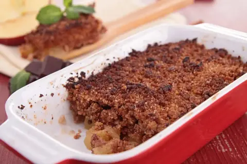 Crumble cacao appena sfornato in una teglia rossa, con mele e cioccolato a lato.