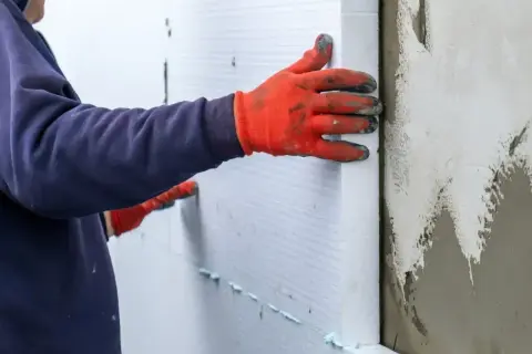 Ręce w pomarańczowych rękawicach przytwierdzają płyty styropianowe do ściany, wykonując ocieplenie stropu styropianem.