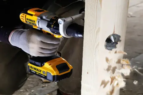 Ręka w rękawicy trzyma wiertarkę DeWalt, która wierci otwór w drewnie. Pokazano, jak włożyć wiertło do wiertarki.