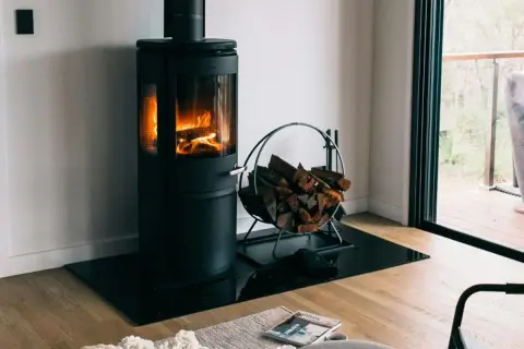 Moderner Holzofen mit brennendem Feuer, daneben ein Korb mit Holzscheiten. Ein gemütlicher Anblick, der an ein Verbot von Holzöfen denken lässt.