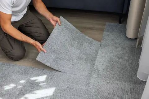 Pose d'une nouvelle moquette pour comment recouvrir un carrelage. Un homme pose une dalle de moquette grise sur un sol en bois.