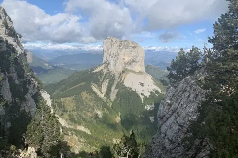 Majestätischer Mont Aiguille ragt aus grünen Tälern empor, umgeben von Felsen und Kiefern unter einem dramatischen Wolkenhimmel.