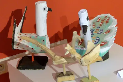 Dwa ptaki z drewna, dzieło Dionizego Purty, z malowanymi piórami i ozdobnymi ciałami, stoją na tle pomarańczowej ściany.