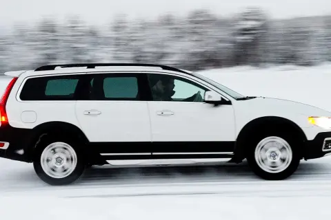Vit Volvo XC70 kör på en snöig väg, med en förare bakom ratten. Bilen är i rörelse, vilket skapar en suddig bakgrund av träd.