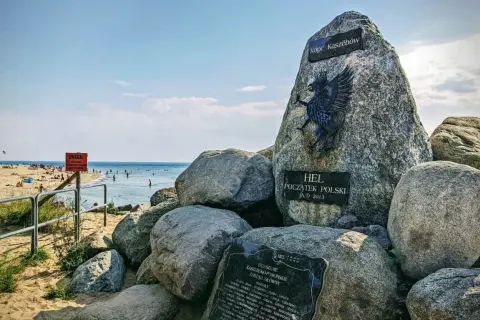 Pomnik "Początek Polski" na plaży na cyplu helskim. W tle widać morze i plażowiczów.