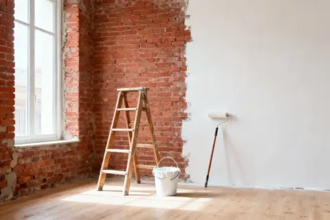 Una habitación con paredes de ladrillo expuesto y una pared parcialmente pintada de blanco. Una escalera de madera, un cubo de pintura y un rodillo esperan para continuar la renovación.