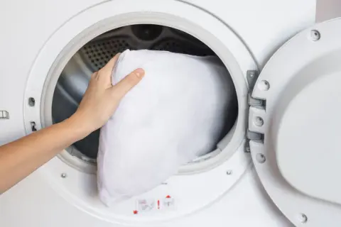 A hand places a white pillow into a washing machine, demonstrating how to wash pillows in the washing machine for a fresh, clean sleep.