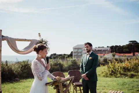 La mariée, en robe blanche, parle au micro lors de la trame cérémonie laïque. Le marié, en costume vert, l'écoute attentivement.