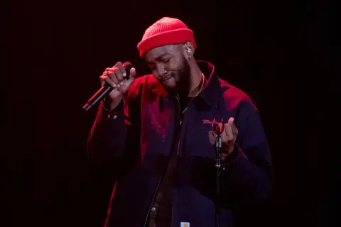 Ant Clemons, wearing a red beanie and dark jacket, sings passionately into a microphone on a dimly lit stage.