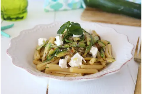 Pasta fredda con zucchine grigliate e cubetti di formaggio fresco, guarnita con erbe aromatiche. Un piatto estivo leggero e gustoso.