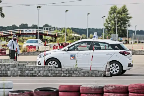 Instruktor kieruje białym samochodem z literą "L" na dachu podczas kursu doszkalającego dla młodych kierowców na placu manewrowym.