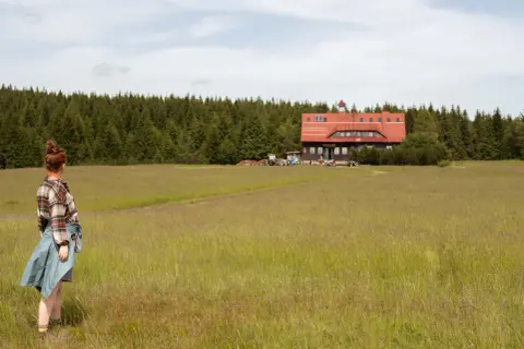 Dziewczyna w kratę podziwia krajobraz w Górach Izerskich. W oddali schronisko i las.