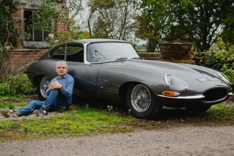 Man sitter vid en grå Jaguar E-Type i lantlig miljö.