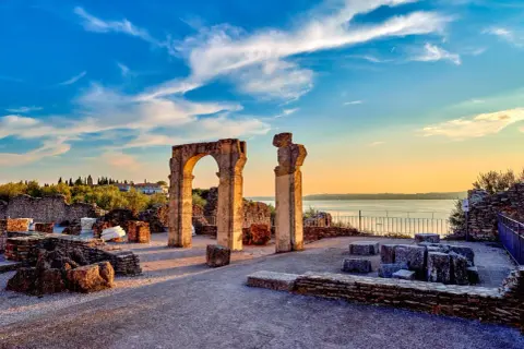Resti archeologici delle Grotte di Catullo con un arco in pietra e colonne, sullo sfondo il lago e un cielo azzurro al tramonto.
