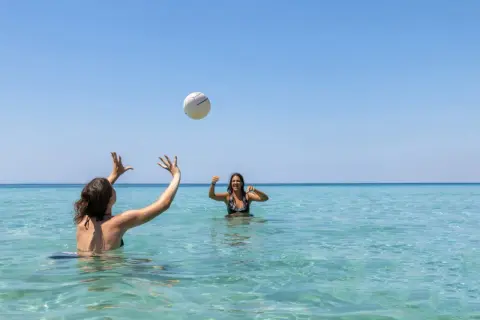 Dos amigas juegan voleibol en el mar turquesa de la playa América, bajo un cielo azul despejado.