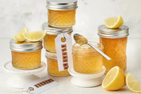 Gläser mit leuchtend gelber Zitronenmarmelade, garniert mit frischen Zitronenspalten. Ein Löffel mit Marmelade zeigt die köstliche Konsistenz, perfekt für ein Zitronenmarmelade Rezept.