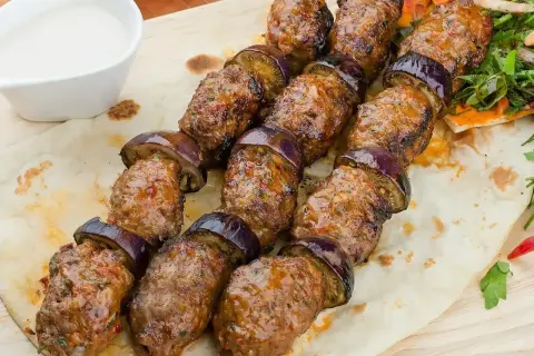 Kofta kebab z bakłażanem na patyku, podany na chlebku z sosem.